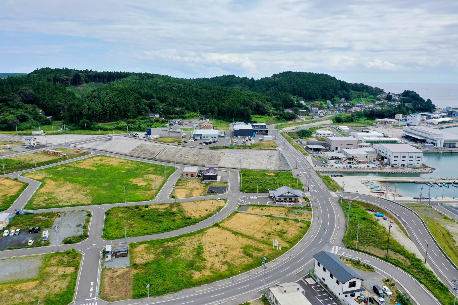 道路が広く区画整理された土地と緑の山々が背景にあり、海岸線に沿って建物や港が並んでいる様子の写真