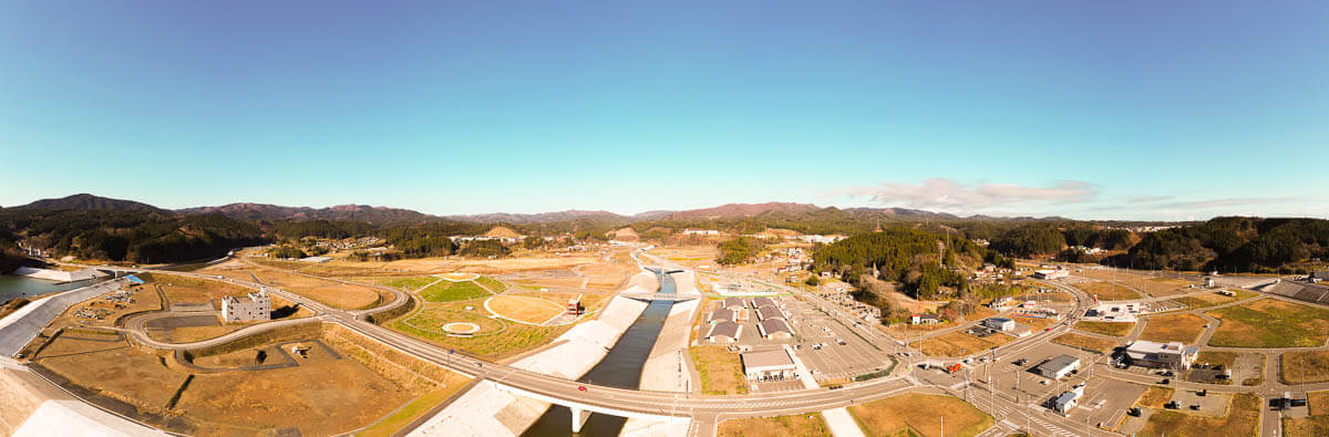 中央には大きな河川と、それを護る広大な防潮堤が整備されており、その上を道路や橋が通っていて河川沿いの両側には造成地が広がり、特に右手には広い駐車場と商業施設のような建物群が見える写真