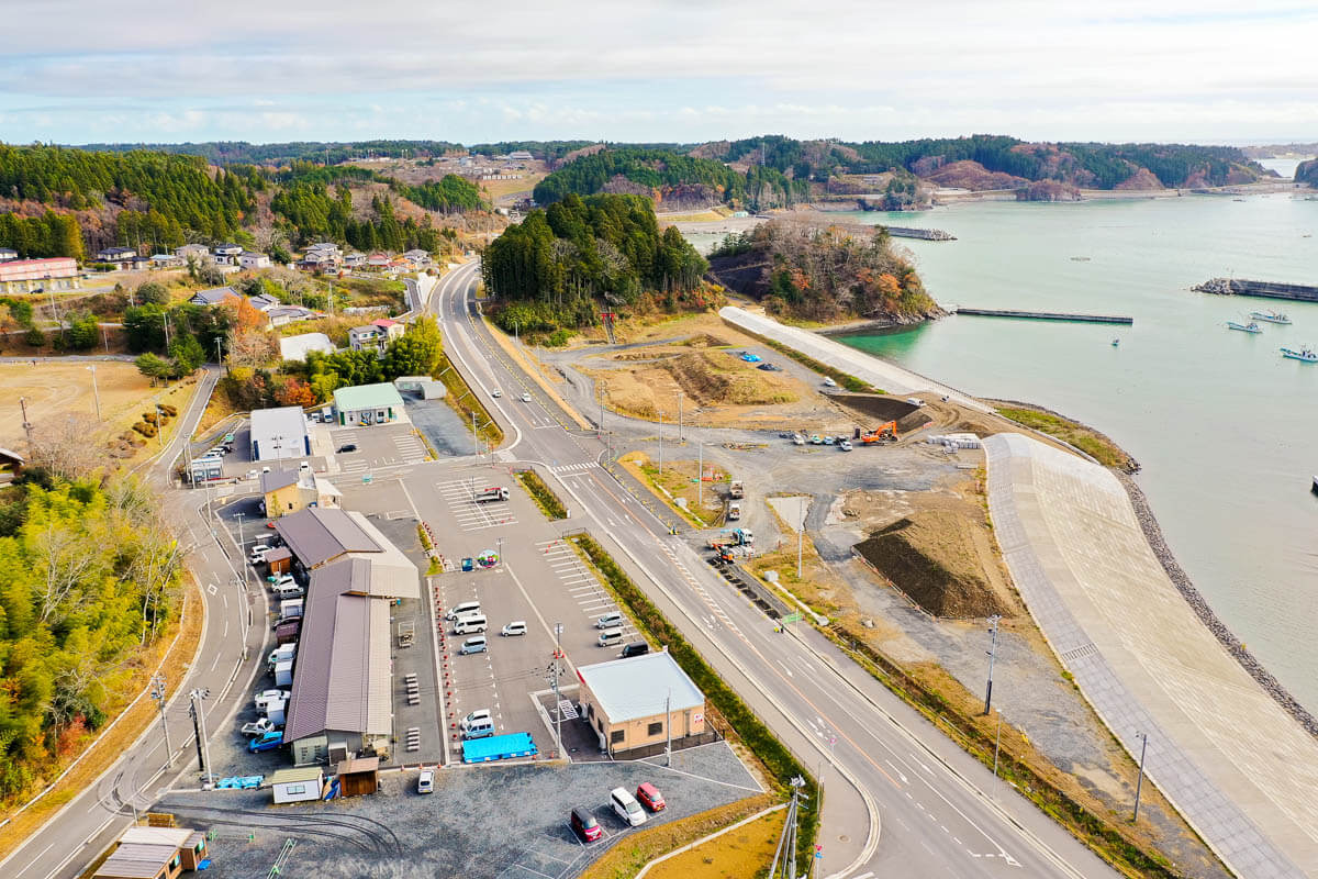 海沿いに白いコンクリート護岸が連続し、津波被害を防ぐ構造となっていて防潮堤の背後には造成地が広がっており、工事車両も見え、大型の駐車場を備えた建物が見える写真道路をはさんで