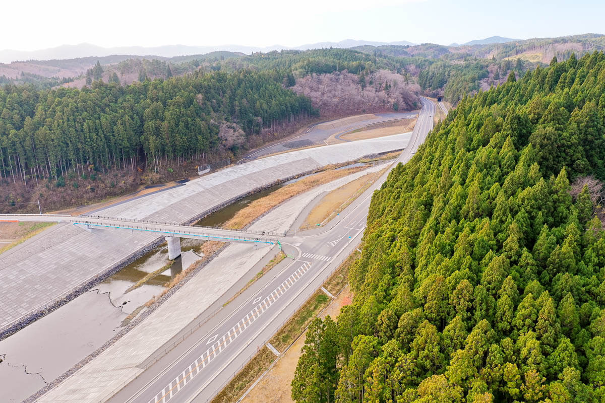 車道は広く、路面も新しく整備され豊かな森林が背後地の自然環境を示し、堤防を跨ぐ形で架けられた歩道橋がみえる写真