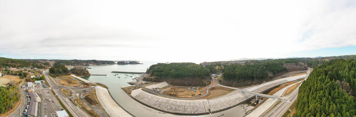 湾口から内陸へ延びる強固な護岸と堤防、道路沿いに商店や住宅が立ち並んでいる写真