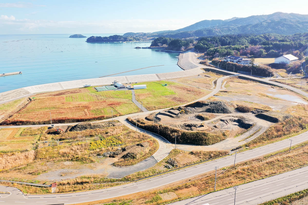手前には造成工事の跡が残り、重機や盛土された区画が見え、道路も新しく整備され、曲線的に走りの奥には農地として利用されているような緑地や畑のような区画があり、小さな施設も確認できる写真
