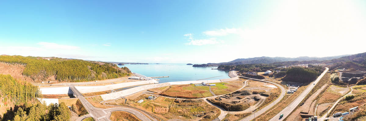 左には森林、中央には港と防潮堤、その内側には区画整備された土地や畑のような緑地が広がっていて、右側には道路や建設途中の造成地が見え、全体的に復興のために整備が進められている地域の様子が伺える写真