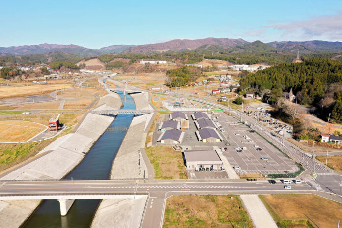 川沿いに整備された堤防と広い駐車場、背後には住宅地と山並みが広がり、全体的に復興の進んだ街の風景が見える写真
