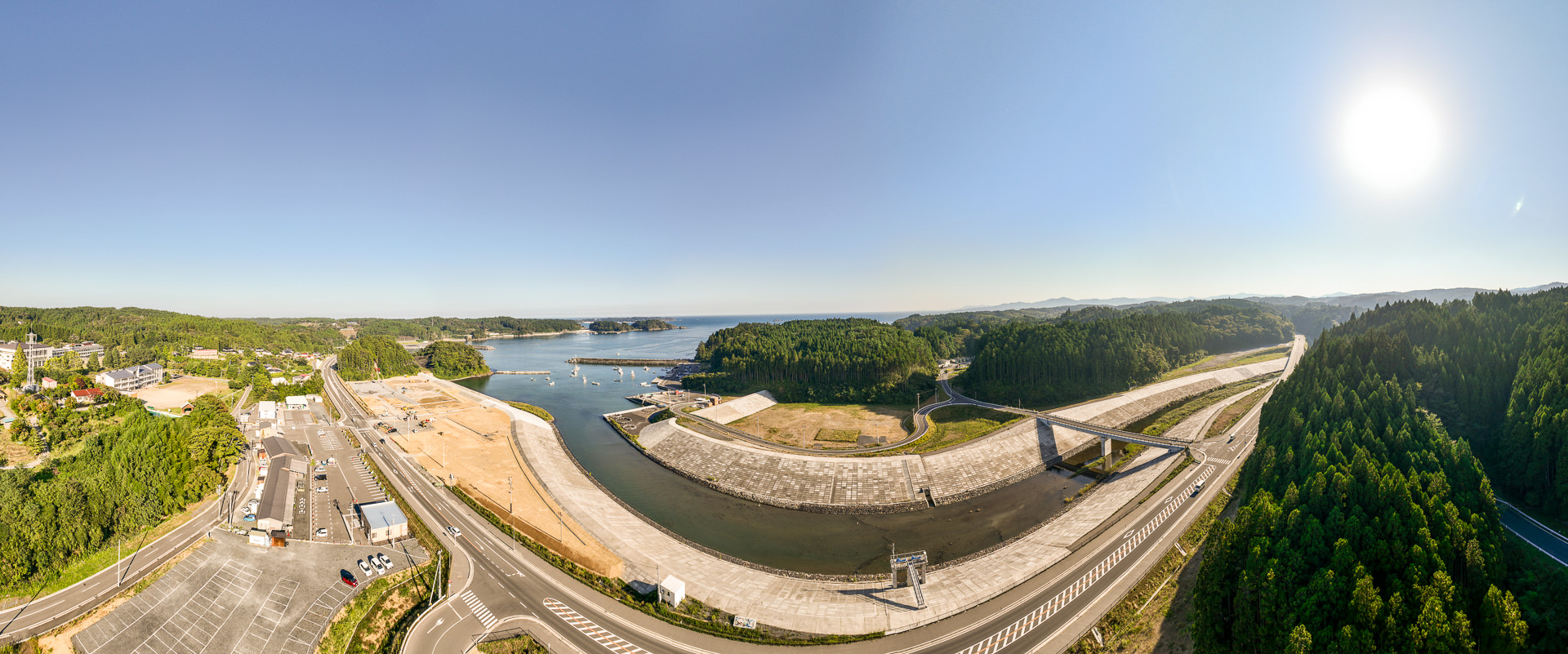 広い河川敷の護岸工事現場と、その横を走る道路、遠くに見える緑の森と海の風景を高台から定点カメラで撮影した写真
