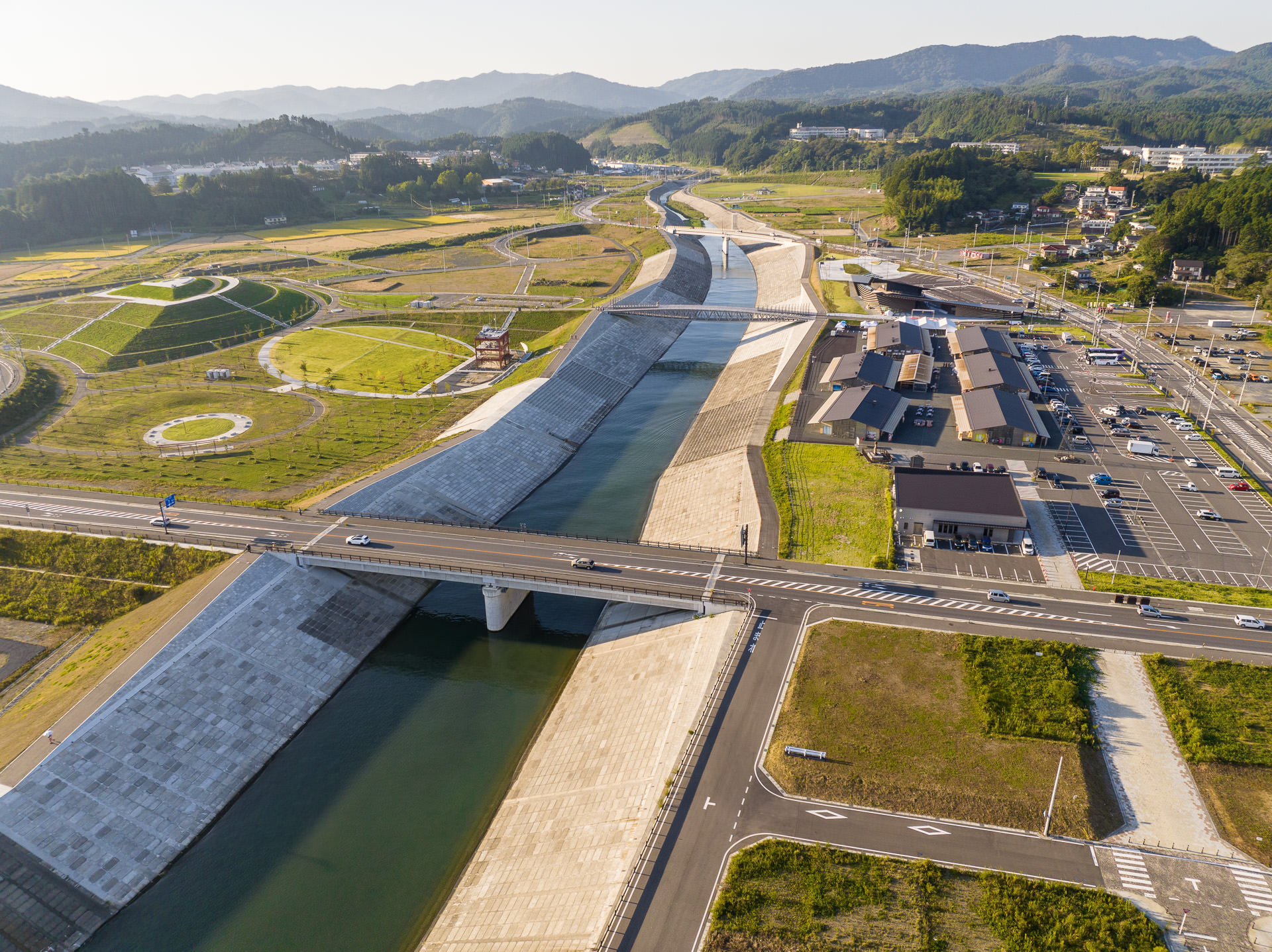 奥へ向かって続く川と橋、その周囲の緑地帯と道路が整然と配置された様子が見える昼間の風景を高台から定点カメラで撮影した写真
