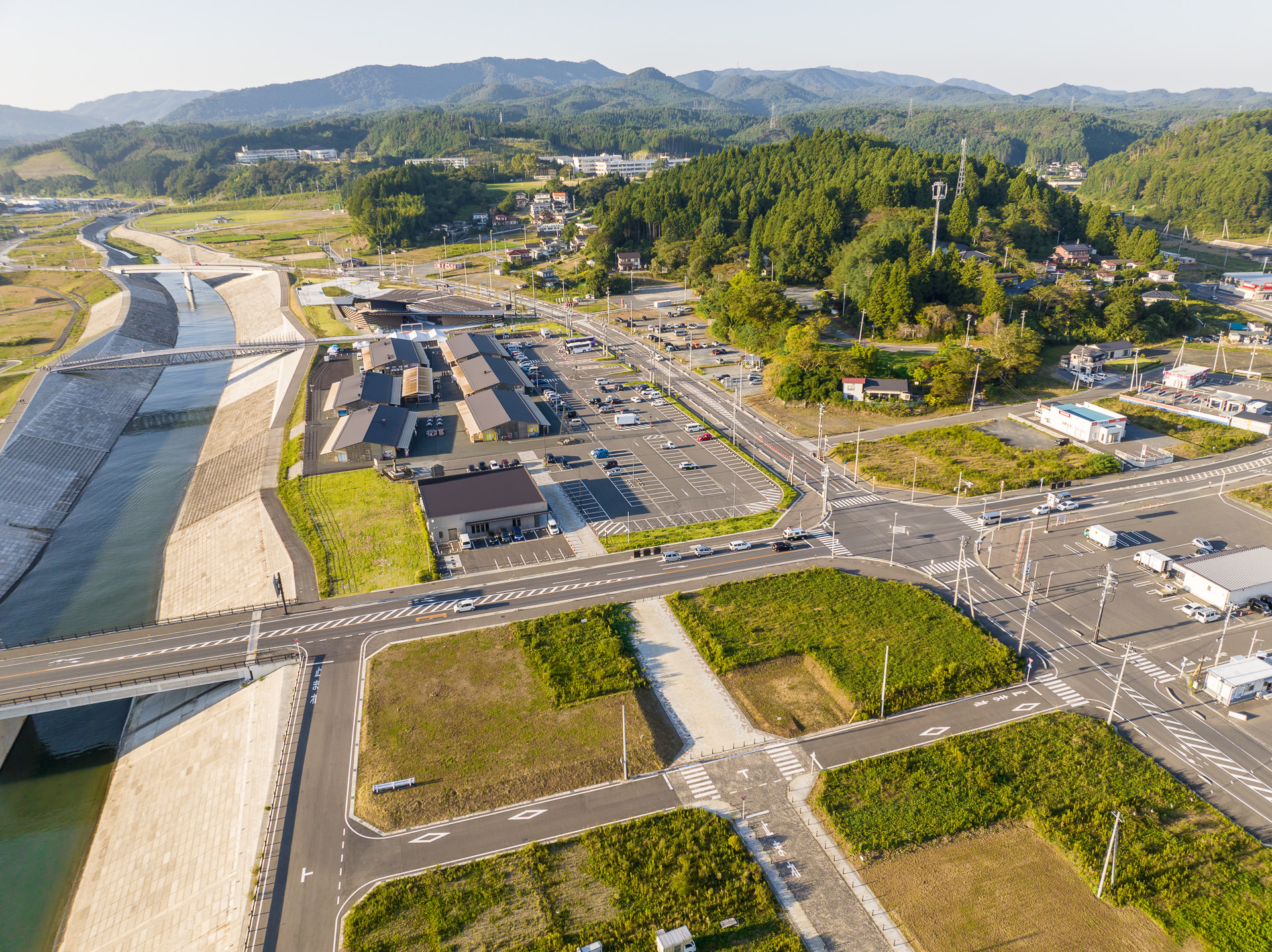 川沿いに建つ施設や広い駐車場、周囲に住宅や森が点在する昼間の風景を高台から定点カメラで撮影した写真