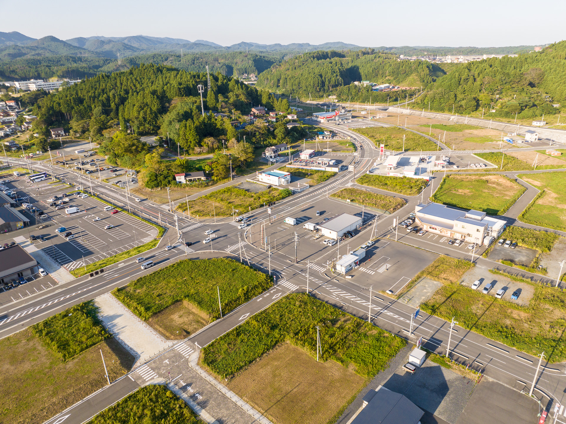 交差点の周囲に住宅や店舗が点在し、周囲を森林に囲まれ、整備された道路や空き地が広がっている町の昼間の風景を高台から定点カメラで撮影した写真