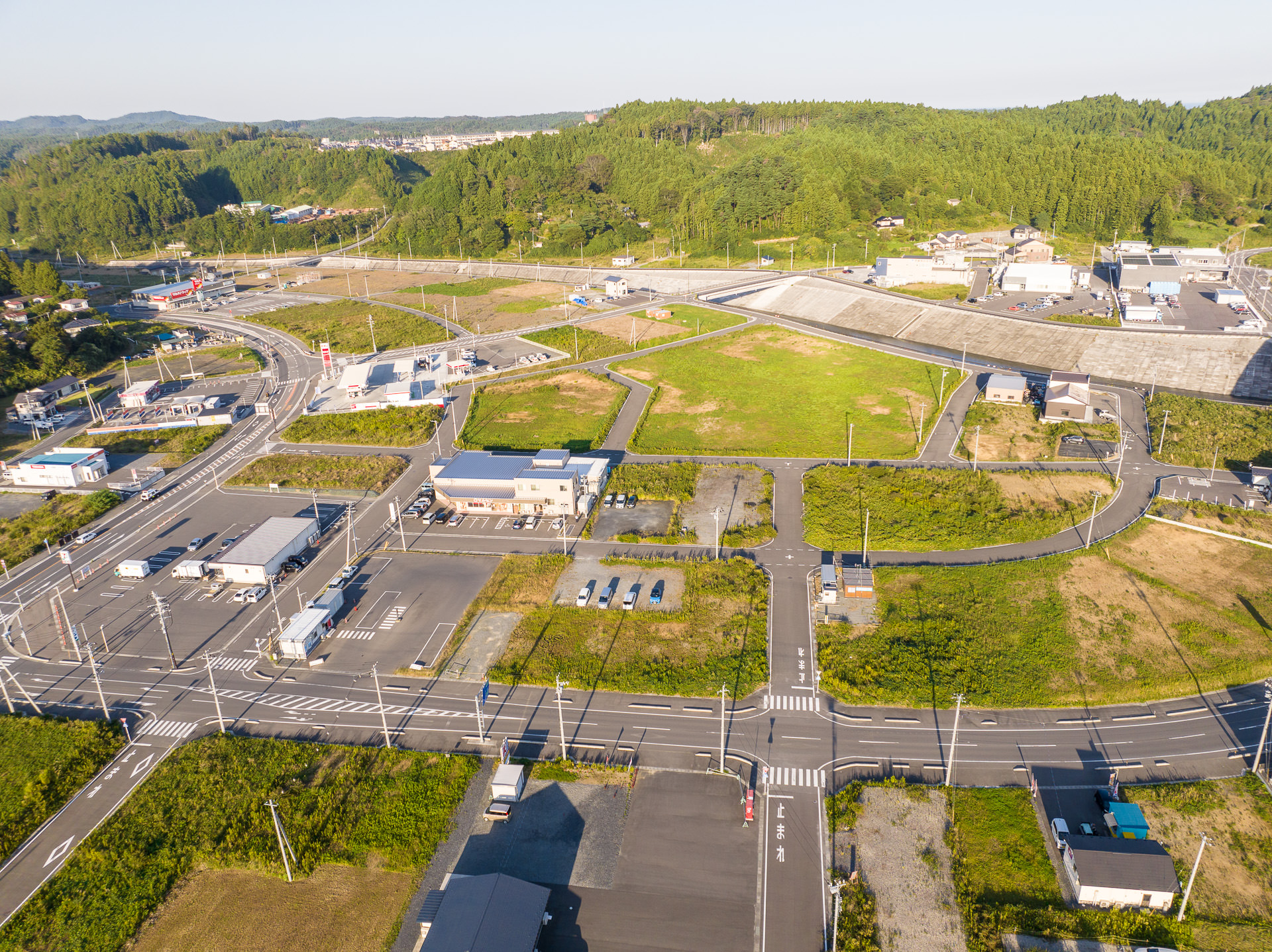 背景に青空が広がる緑の丘陵地帯と整然と区画された道路や建物が見える陸地の風景を高台から定点カメラで撮影した写真