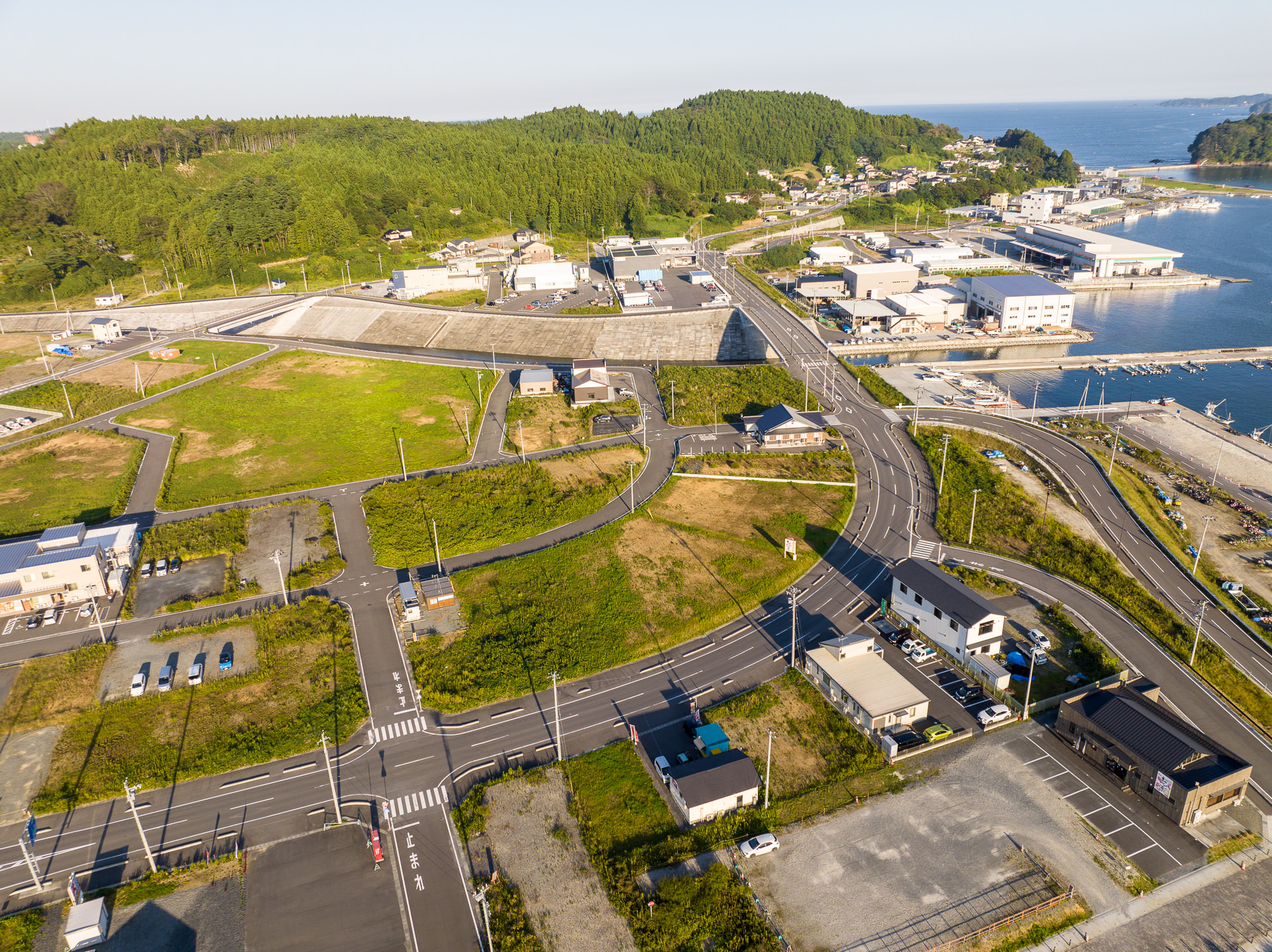 港へ続く道路沿いに建物が点在し、奥には緑の山々と海が見える風景を高台から定点カメラで撮影した写真