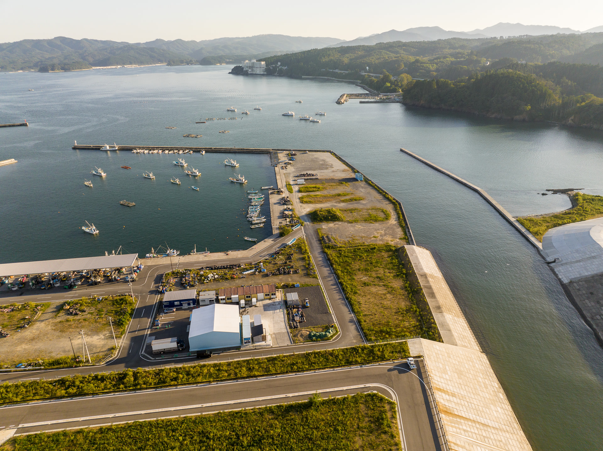 防波堤で囲まれた港とその周辺の陸地、複数の漁船が停泊し、山並みが遠景にある海岸地域の風景を高台から定点カメラで撮影した写真