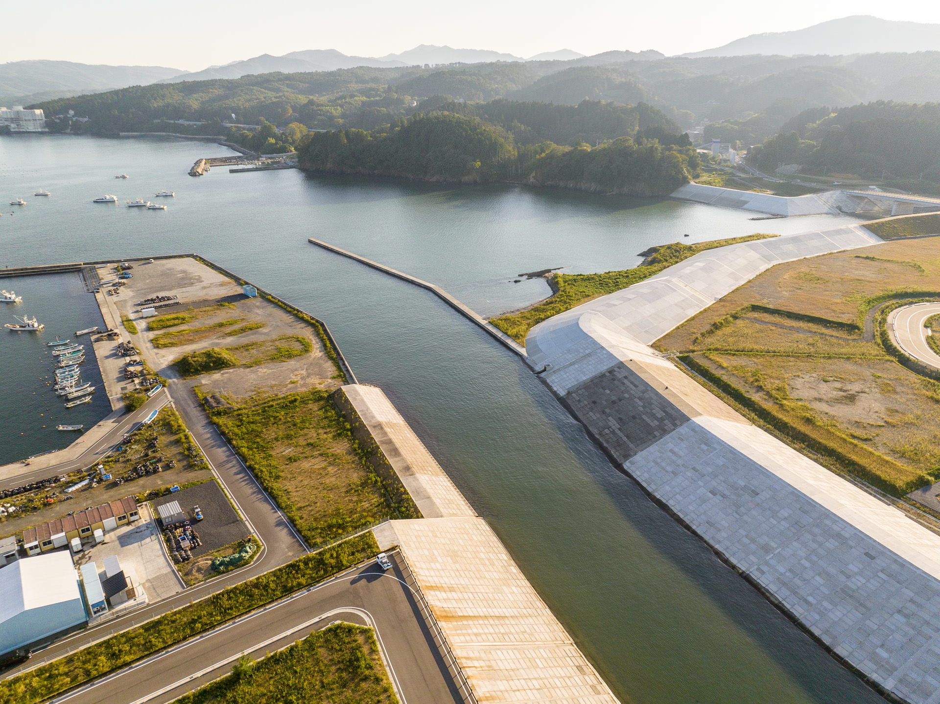 港の入り口の水路とコンクリート護岸の両岸の防潮堤、岸辺に並ぶ漁船や倉庫が見える風景を高台から定点カメラで撮影した写真