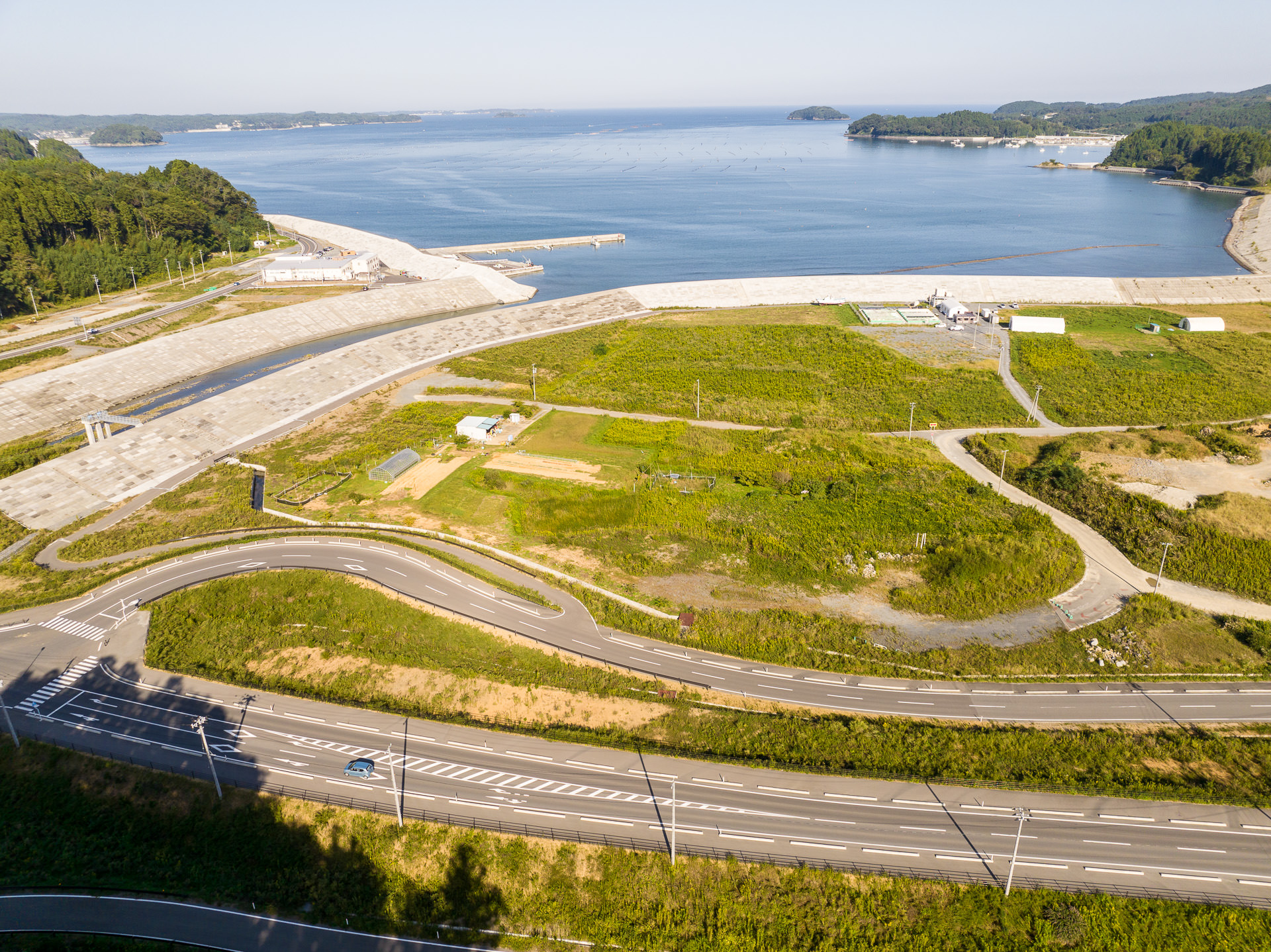 手前にまっすぐな道路とゆるやかにカーブした道路があり、道路と湾の間に点在する建物と緑が広がる土地が広がり、海岸に接する護岸が長く続いて整備されている昼間の風景を高台から定点カメラで撮影した写真