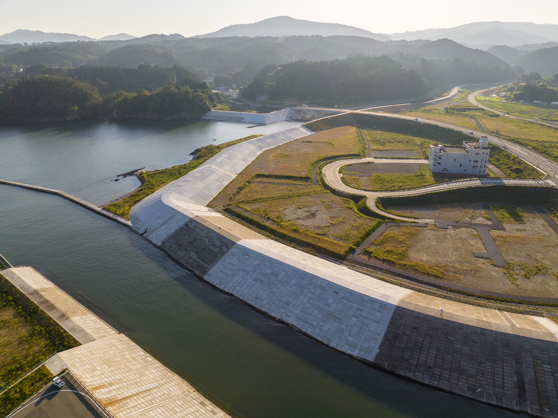 防潮堤の曲線と内部の草地、中央に立つ数階建ての白い建物が、遠くの山々とともに写された風景を高台から定点カメラで撮影した写真