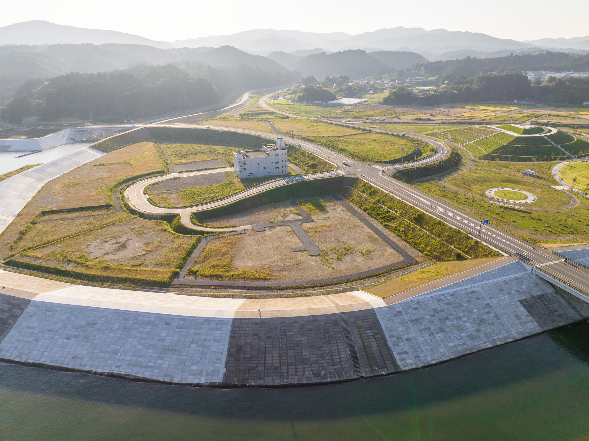 防潮堤に囲まれた草地の中に、道路が取り巻く数階建ての白い建物が建っている様子が、遠くの山々を背景にした風景を高台から定点カメラで撮影した写真
