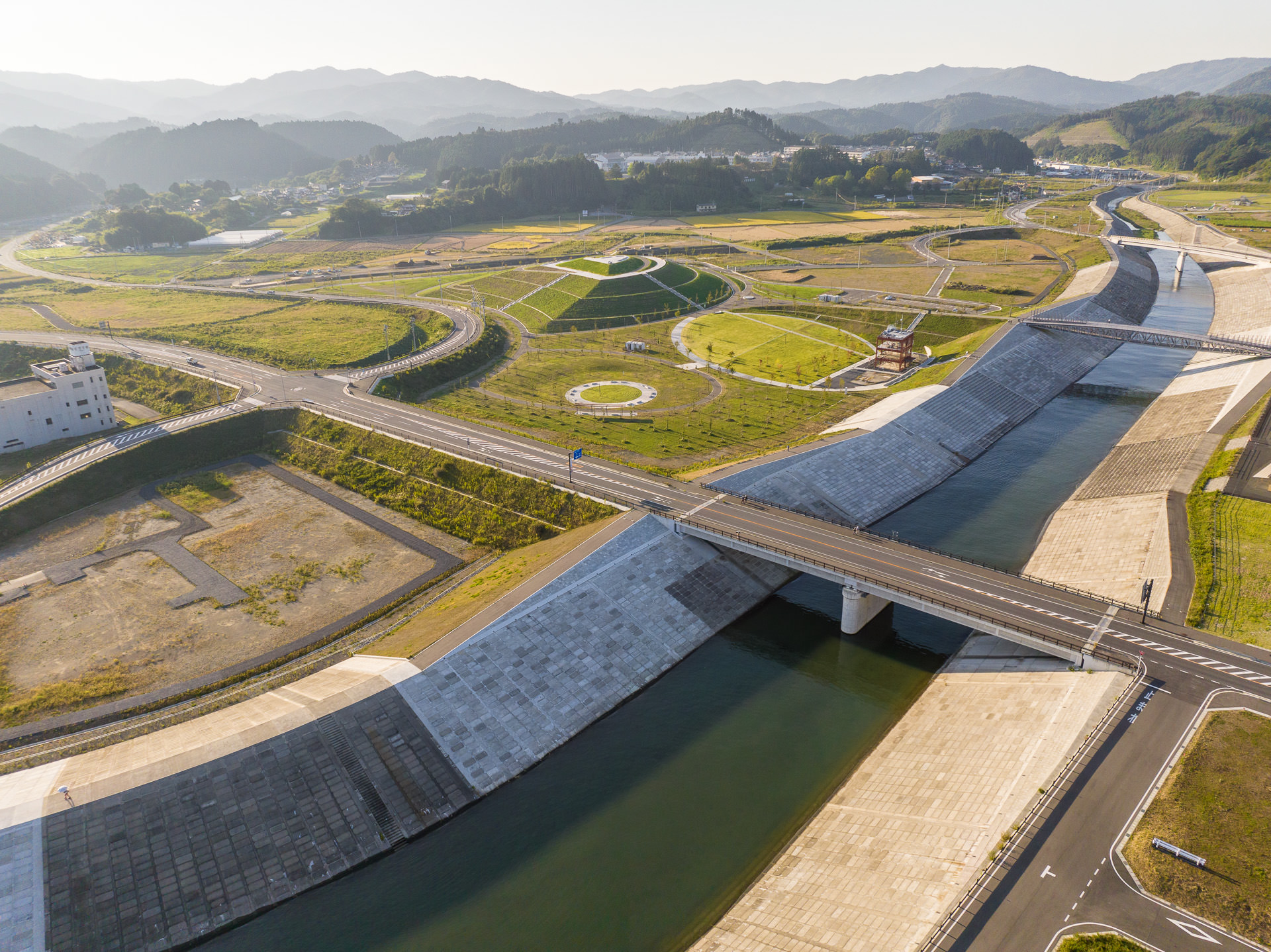 川に架かる橋とその周囲に広がる緑地帯、道路や麓に集落がある山々が遠くに見える昼間の風景を高台から定点カメラで撮影した写真
