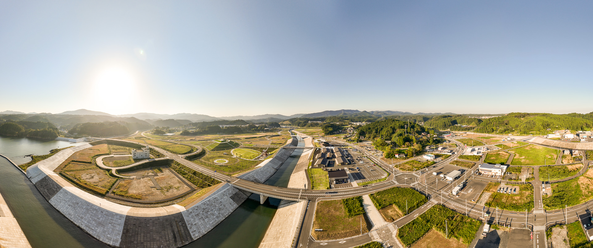 中央に湾へ続くまっすぐな川の両岸に整備された土手と橋が見え、周囲には緑地や道路が広がる昼間の風景を高台から定点カメラで撮影した写真