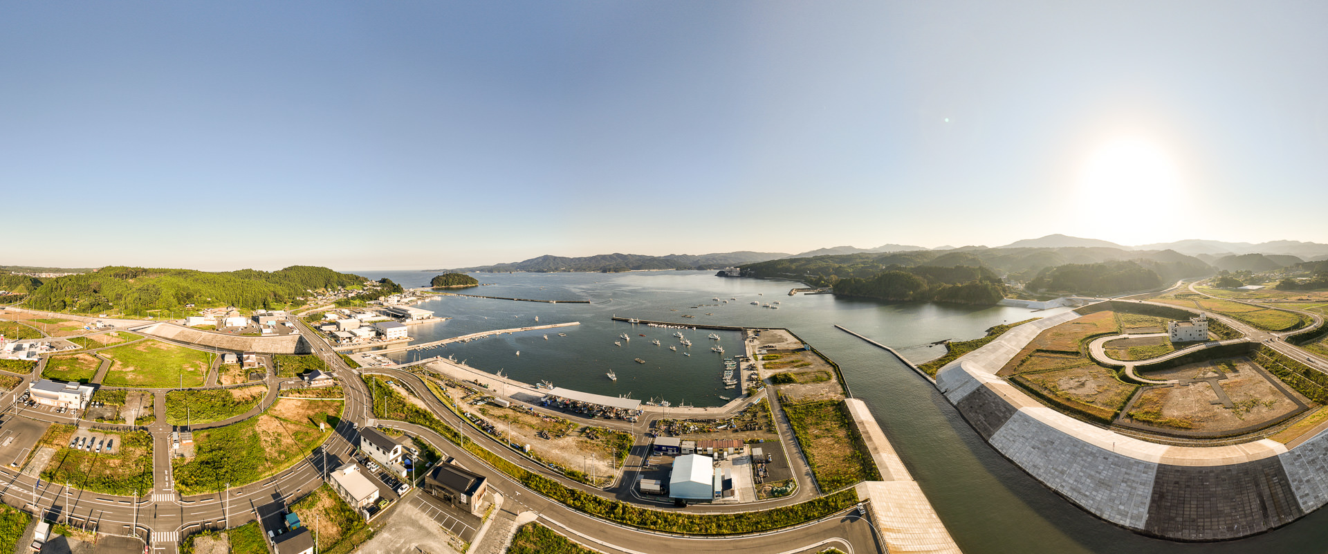 港の全景が広く映され、多数の船が停泊し、防波堤や岸壁が整備された海辺の風景を高台から定点カメラで撮影した写真