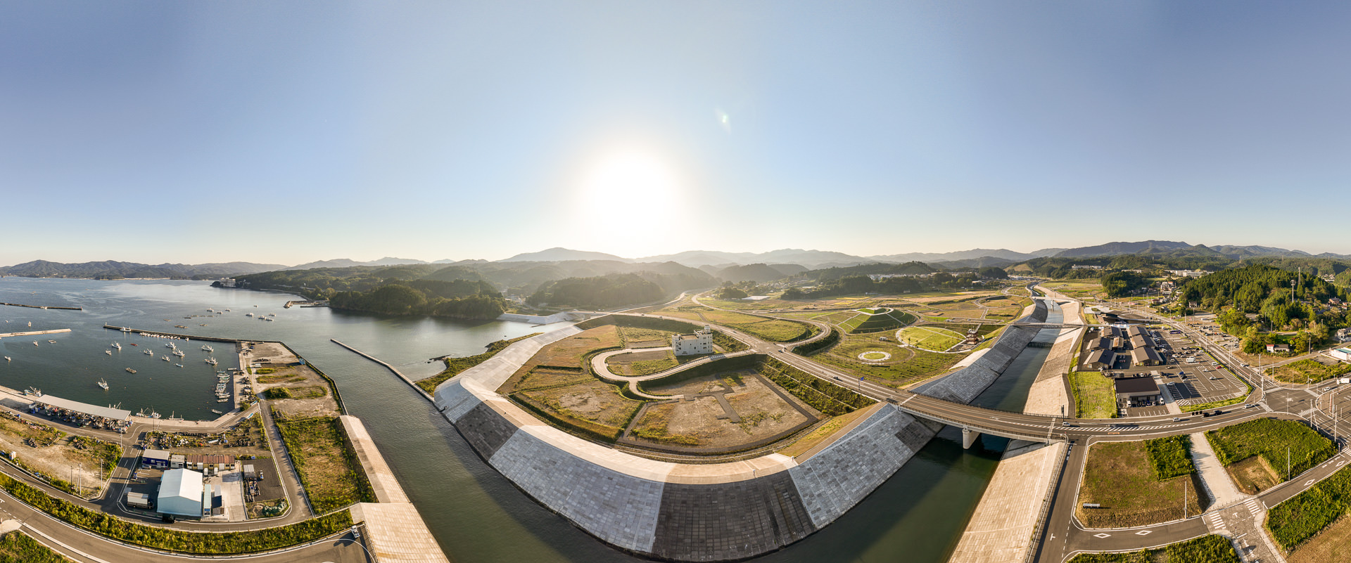 港の入り口から内陸に向けて整備された広大な防潮堤と、その中央にある数階建ての建物の周りに緑の広場が広がっている風景を高台から定点カメラで撮影した写真