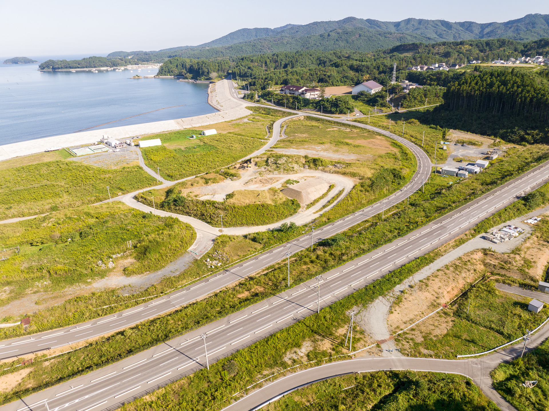 海岸線に沿ってカーブした道路と緑の広場があり、その背後には山並みと住宅地が広がる昼間の風景を高台から定点カメラで撮影した写真