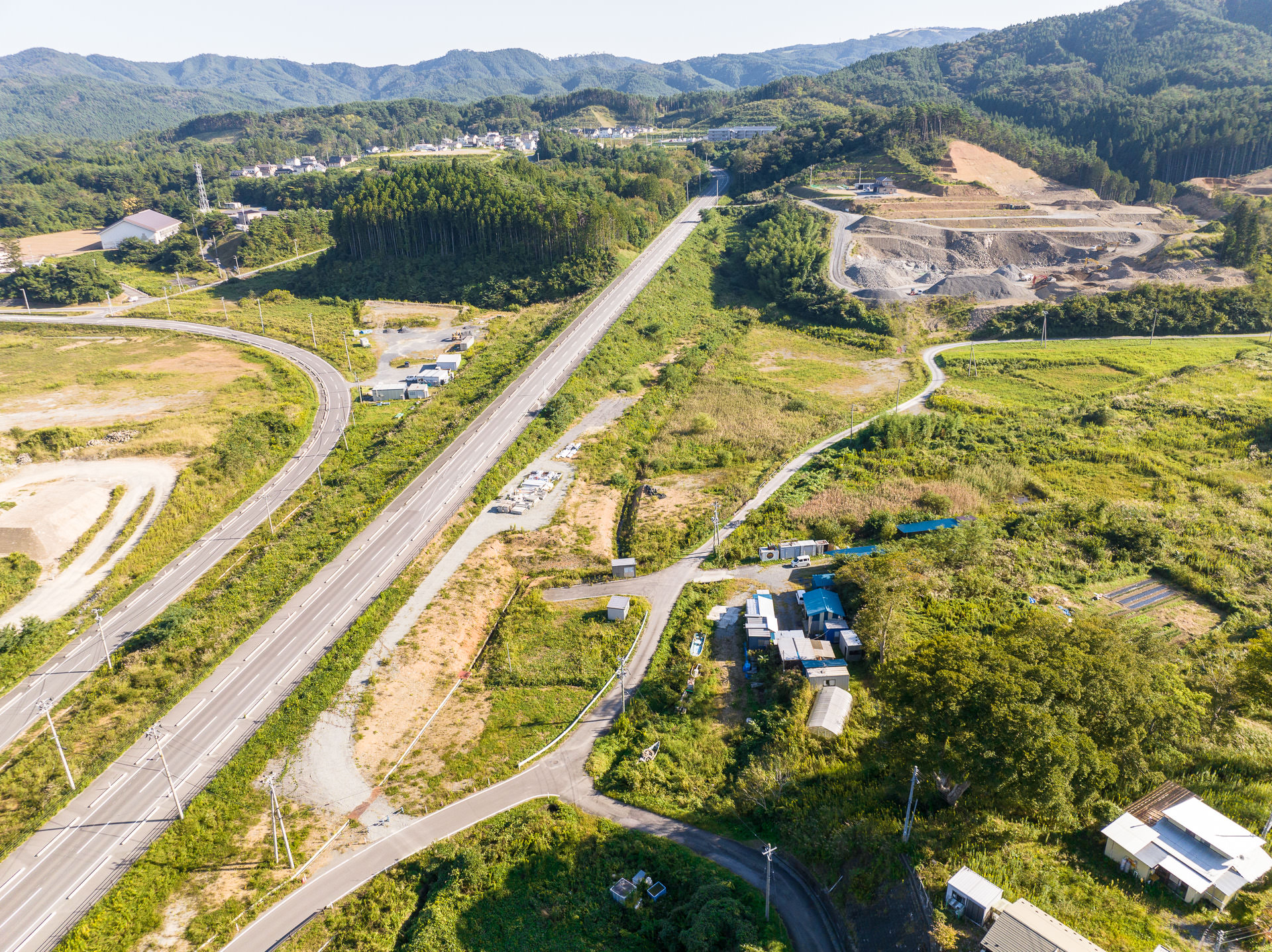 奥まで真っすぐ続く道路と曲がりくねった山間の道が見え、緑の中に散在する住宅や施設、遠くに山が連なる昼間の風景を高台から定点カメラで撮影した写真