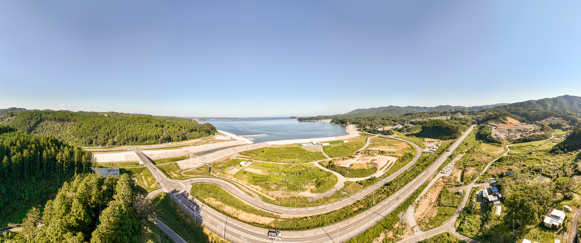 緑の丘と海に囲まれた広大な土地があり、湾内に小さな島が点在し、湾の入口に護岸が整備された昼間の風景を高台から定点カメラで撮影した写真