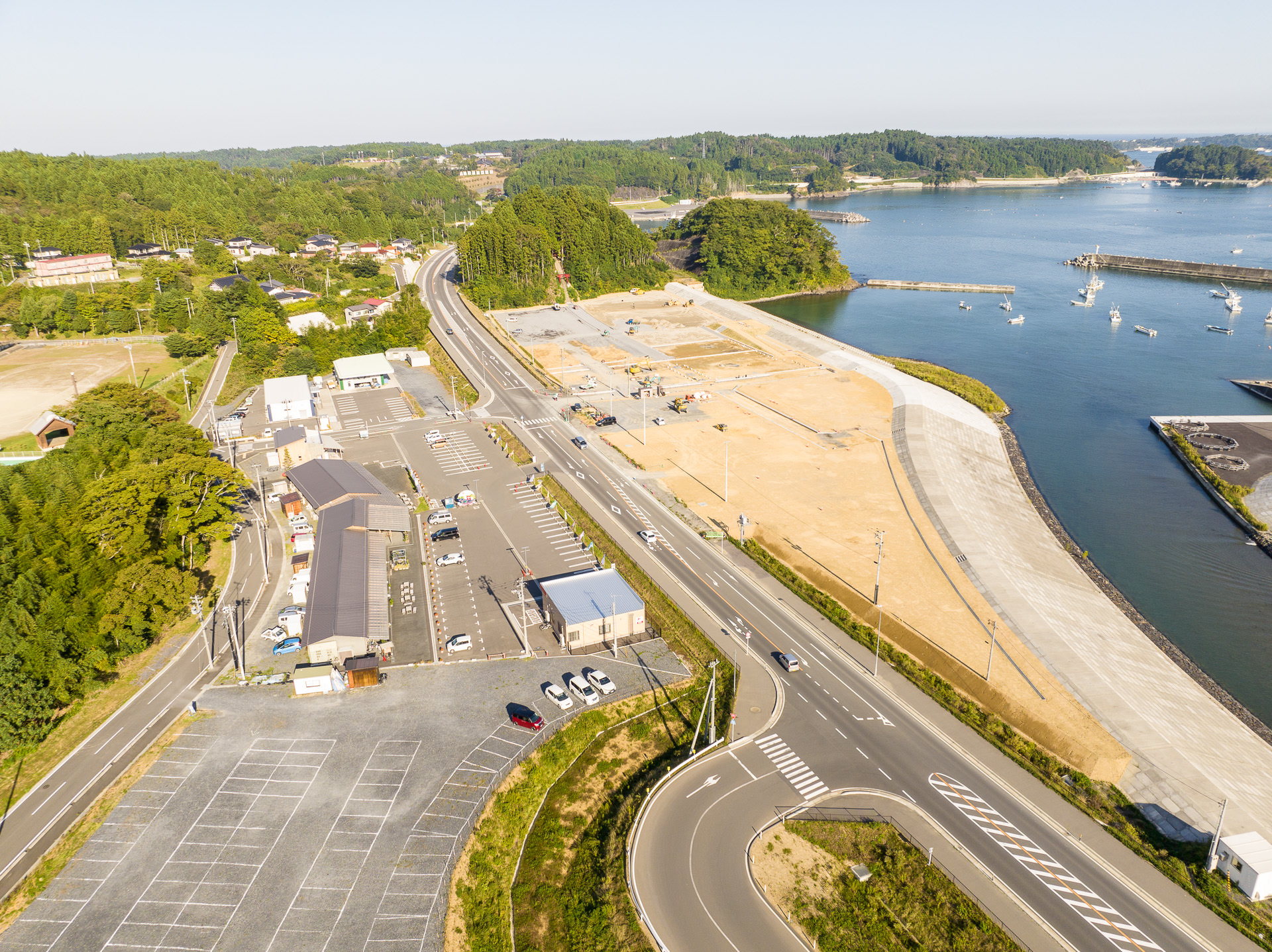河川敷の護岸工事現場に隣接する建物と駐車場、奥まで続く道路、奥には木々と海が見える風景を高台から定点カメラで撮影した写真