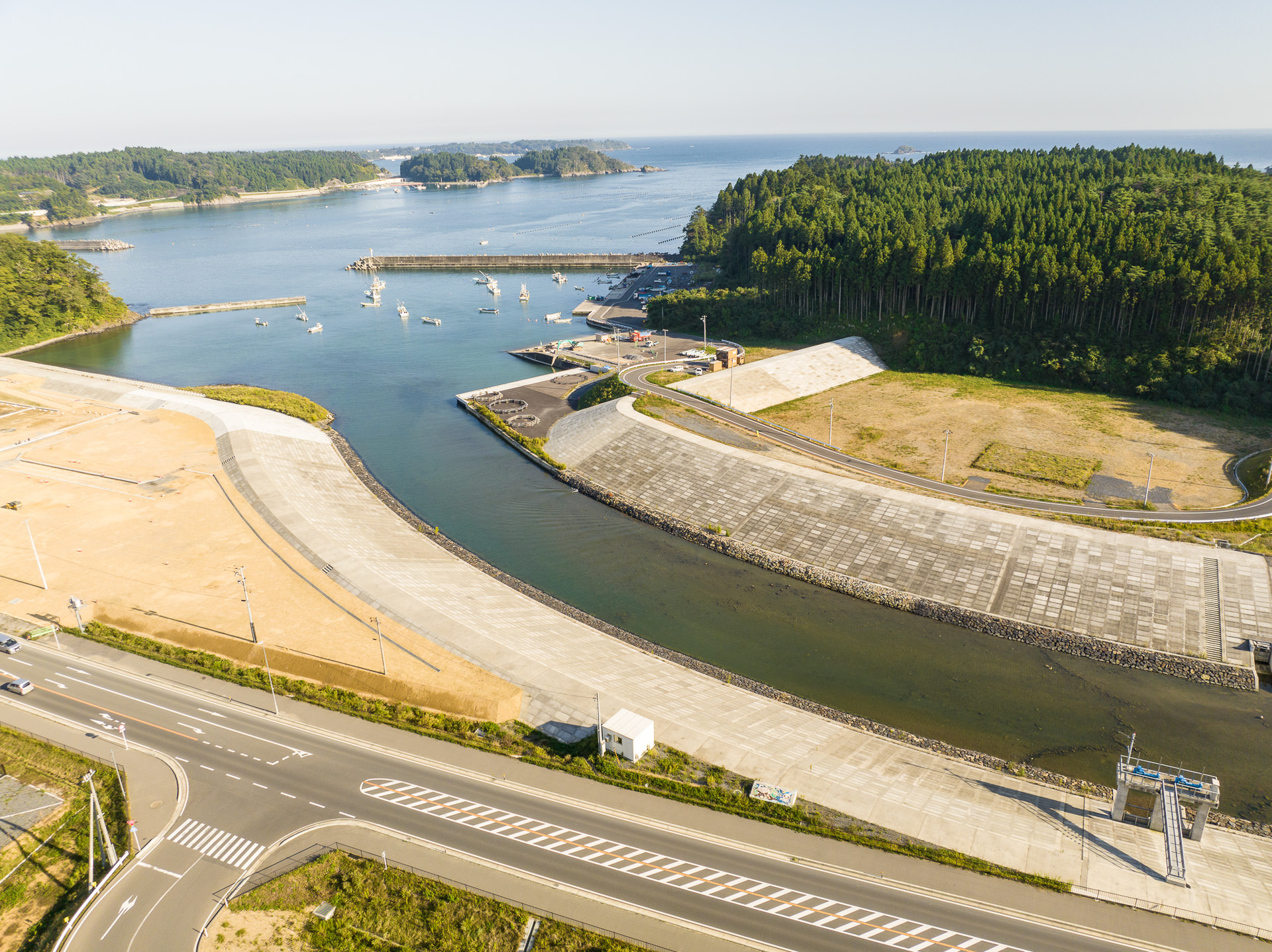 河川敷の護岸工事現場の広いコンクリート護岸と、川を挟んで対岸の緑の森と海が見える風景を高台から定点カメラで撮影した写真