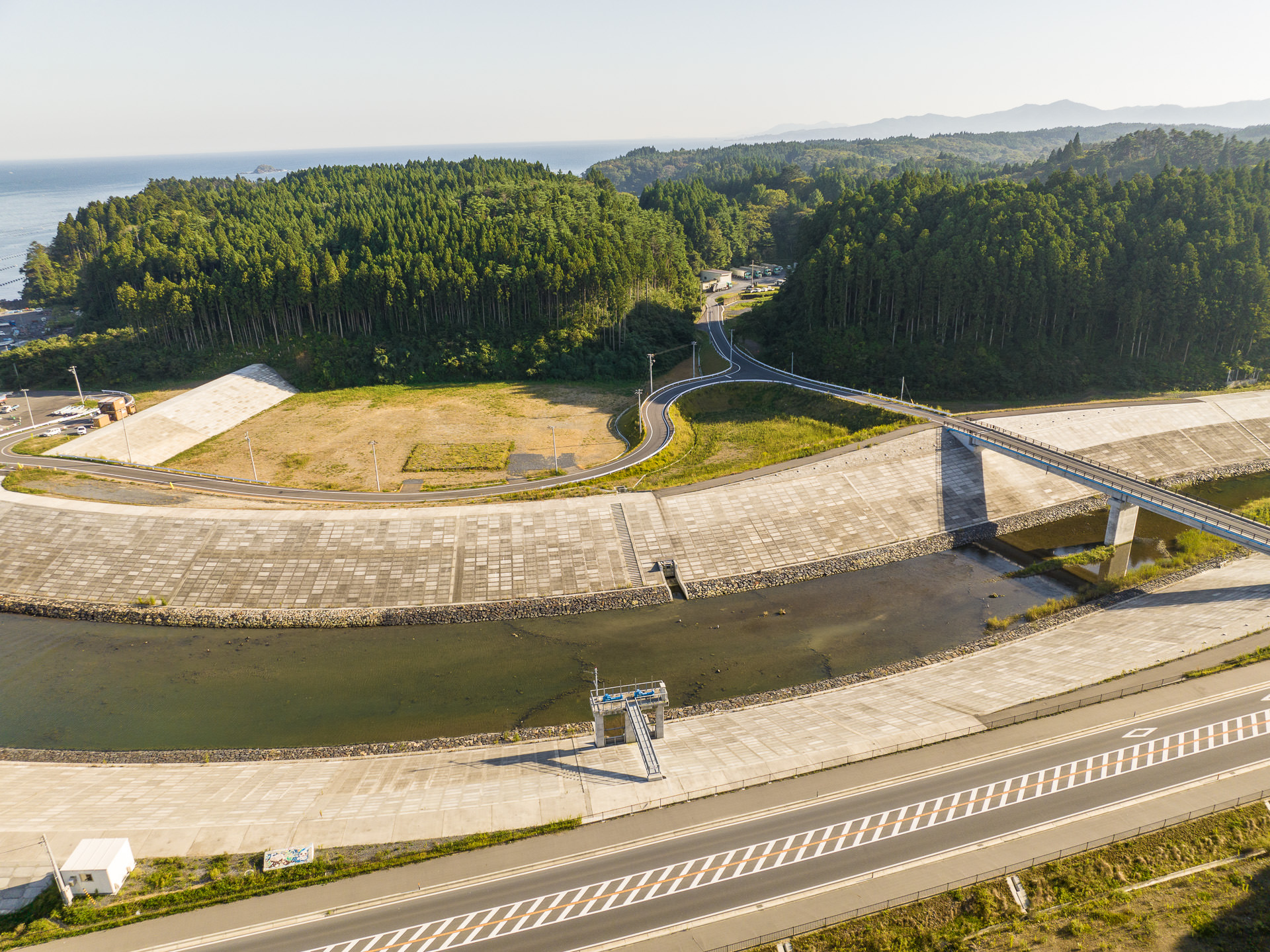 河川敷の護岸工事現場の舗装された堤防と、その上に架かる橋、背景に広がる森林の風景を高台から定点カメラで撮影した写真
