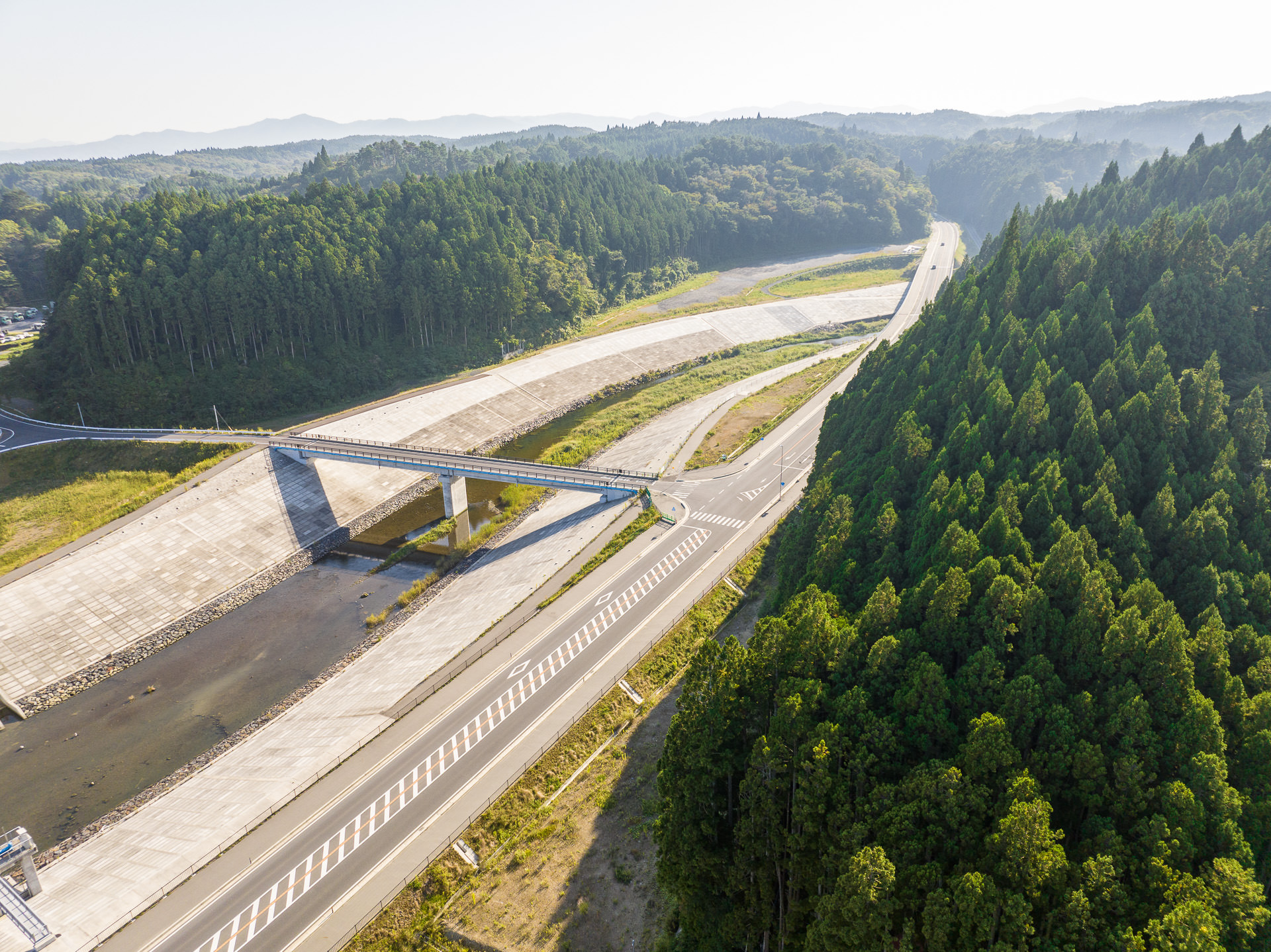 川の護岸工事が施された堤防と橋、横に並ぶ道路と隣接する緑豊かな森林の風景を高台から定点カメラで撮影した写真