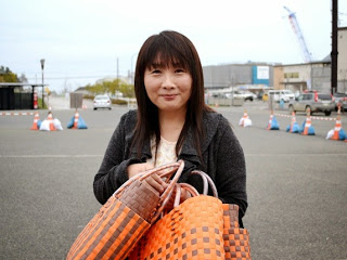 屋外の駐車場で女性が笑顔でカメラを見つめながら、オレンジと茶色のチェック柄のかごバッグを複数抱えている写真