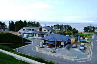 湾曲した道路に沿って建てられた住宅と、周囲に停車している車、背景に広がる青い海と緑の木々が調和している様子が写っている住宅街の写真