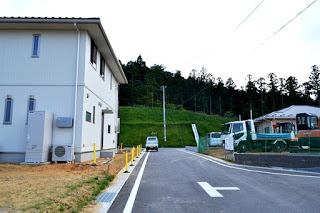 左側に白い二階建ての住宅が建ち、中央には舗装された道路が伸び、右側には工事用の車両や機材が並び、背景には緑の木々が茂る丘が広がっている写真