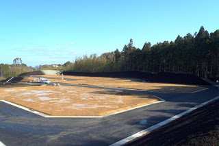 建物がまだ建っていない広い敷地が舗装と土のままで広がり、背景には木々が並び、空が晴れている開発前の土地の写真