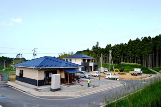 青い屋根の住宅が2棟建てられている造成地に、複数の車両と建設機材が並び、作業員が作業をしている様子が写っている住宅建設現場の写真