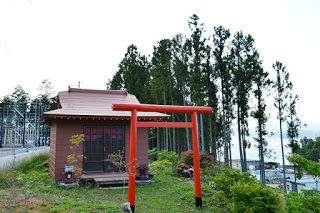 周囲は手入れされた植栽や草花に囲まれ、背景には背の高い杉の木々が立ち並び、赤い鳥居の奥に小さな社が写っている写真