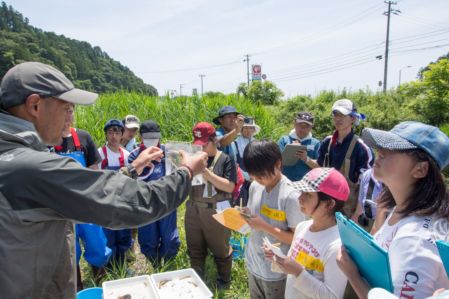 屋外の草地に複数の大人や子供たちが集まり、小さな魚が入っている透明の容器を持って説明している男性の話を、子供たちが熱心にノートやクリップボードを持ってメモをとっている写真