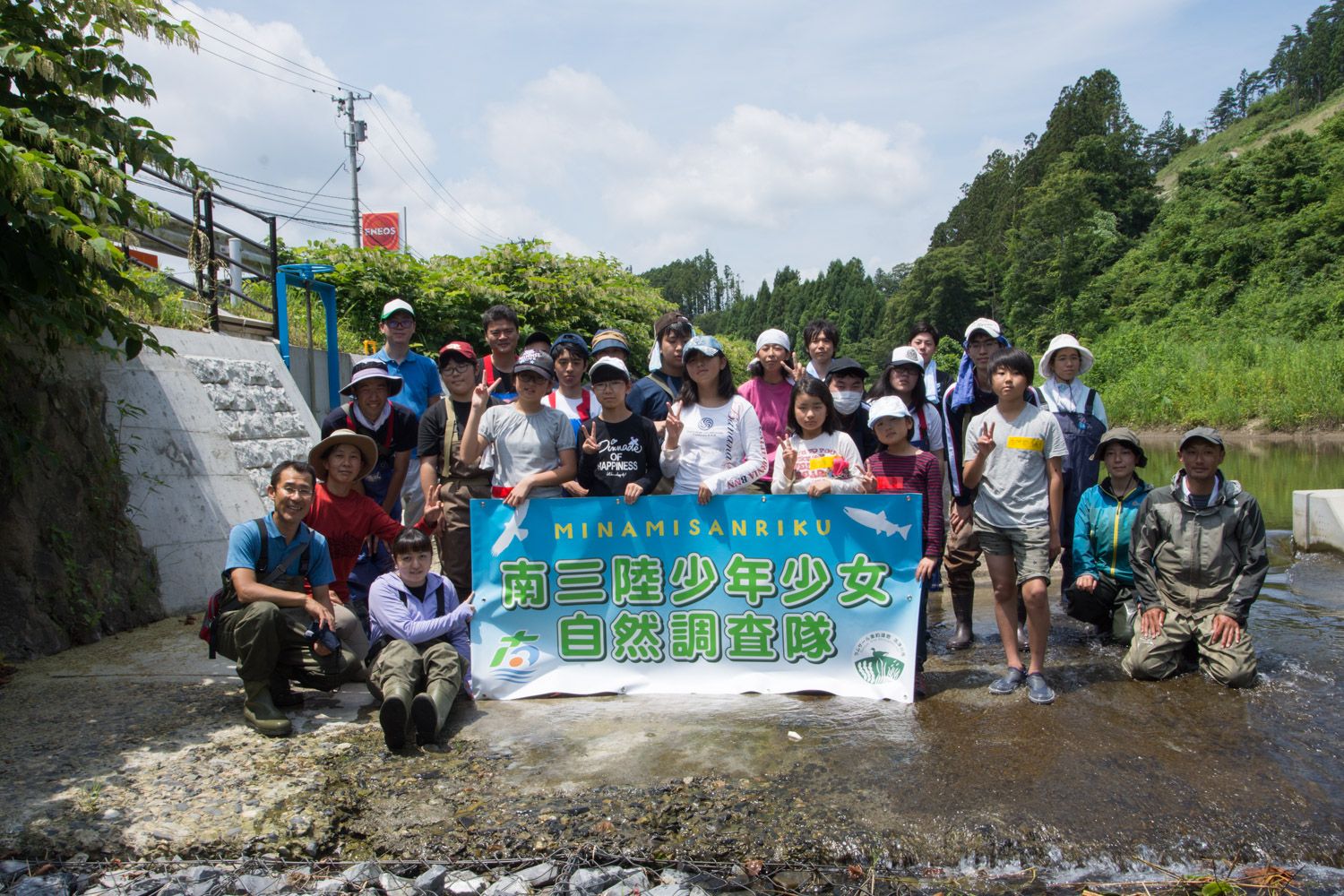 屋外の小さな水辺の中で、「MINAMISANRIKU 南三陸少年少女自然調査隊」と書かれた横断幕を持っている大人や、ピースサインをする子供の参加者が並んでいる集合写真