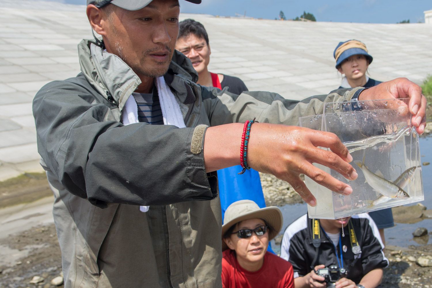 後ろで他の人々が見ているなか、水と魚の入った透明な容器を手に持っている男性が話をしている写真