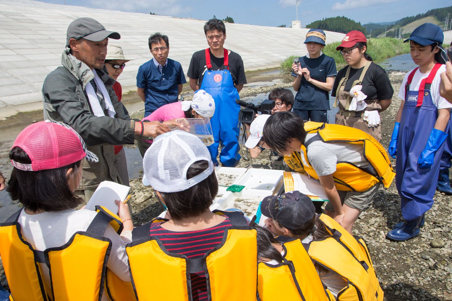 水がない川の中で、複数の人物がライフジャケットや防水のオーバーオール、手袋を着用して集まり、水の入った容器を持って話をしている男性を他の人たちが熱心に聞いている様子が写されている写真