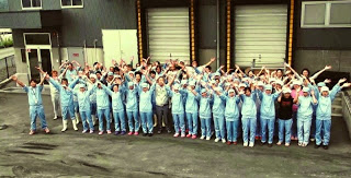 水色の作業服と帽子を着た大勢の人たちが工場のような建物の前で並び、全員で両手を上げて元気よくポーズを取っている集合写真