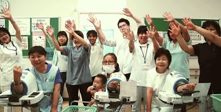 病院のような場所で3人のスタッフが血圧測定器に腕を通し、後方で医療スタッフや看護師たちが笑顔で手を振っている写真