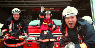 消防車の前で左手に拡声器を持ち、右手を真っすぐ前にあげている消防服を着た消防士を中央に、両端の2人の消防士がホースを持ってポーズを取っている写真