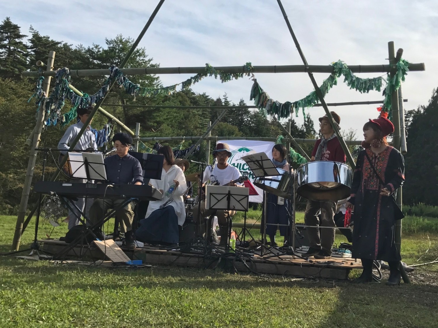 野外の緑豊かな場所に竹で組まれた簡素なステージがあり、そこに7人の演奏者が楽器を演奏している様子が見られ、左から電子ピアノを弾く男性、アコースティックギターの男性、スティールパンを持つ男性、タンバリンを持つ女性、背景にドラムを叩く人やキーボードを持つ人がおり、ステージ上には楽譜台が置かれている写真