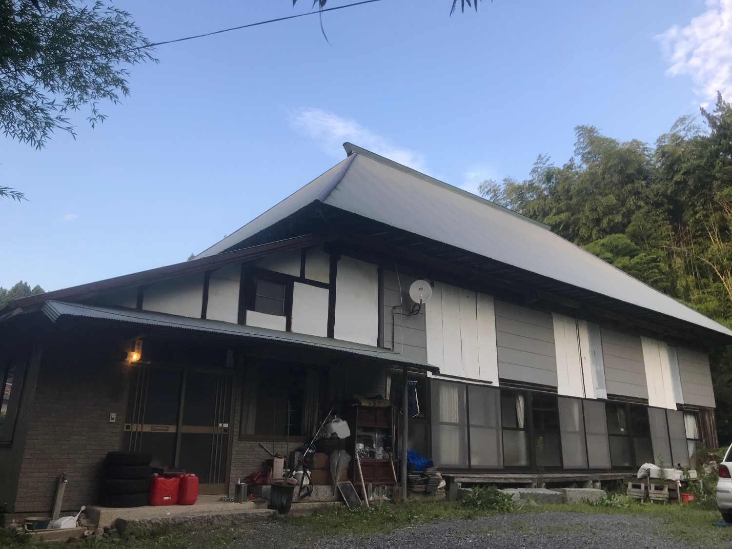 玄関付近にはタイヤや赤いポリタンクが置かれ、建物の外壁は一部が白と灰色の板で覆われている屋根が急勾配で大きく張り出している日本の伝統的な民家の写真