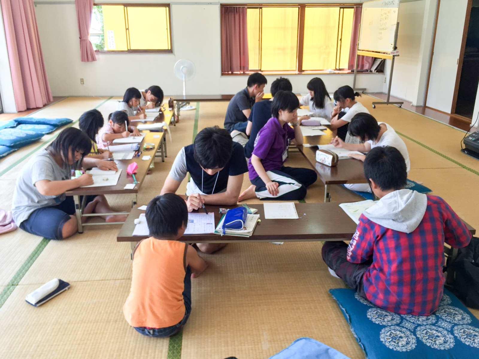 大小の窓がある広い畳の部屋の中で、紙や筆記用具が置かれている口の字型に置かれた長机を囲んで子どもと大人が向かい合って畳に座り、宿題に取り組んでいる様子の写真