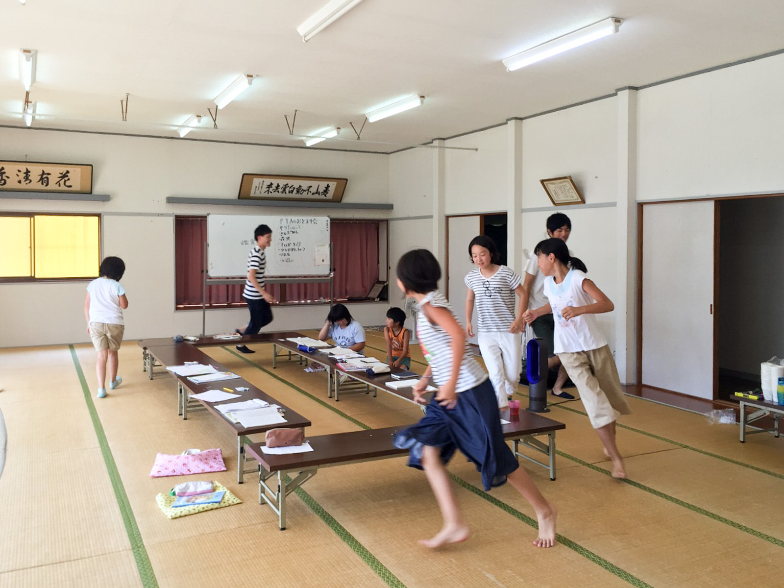 壁には額入りの書道作品が飾られ、文字が書かれたホワイトボードが設置された広い畳の部屋の中で、プリントや筆記用具などが置かれたロの字型に設置された机の周りを子どもと大人が鬼ごっこで走り回ったりして遊んでいる写真