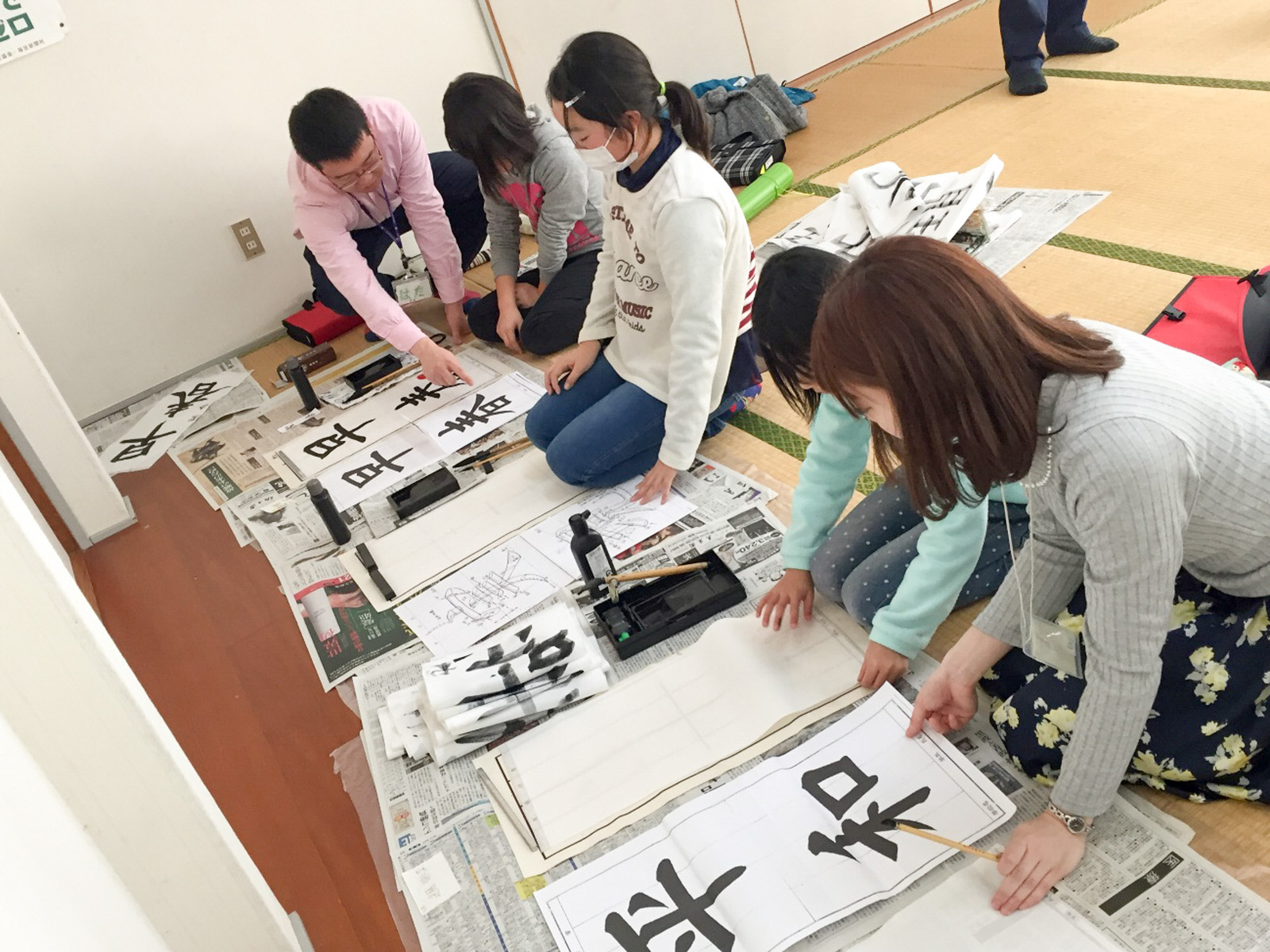畳の上に座り、床に広げた新聞紙の上に半紙と「早春」「平和」のお手本を並べ書道に取り組んでいる子どもたちと、指導をしている男女の写真