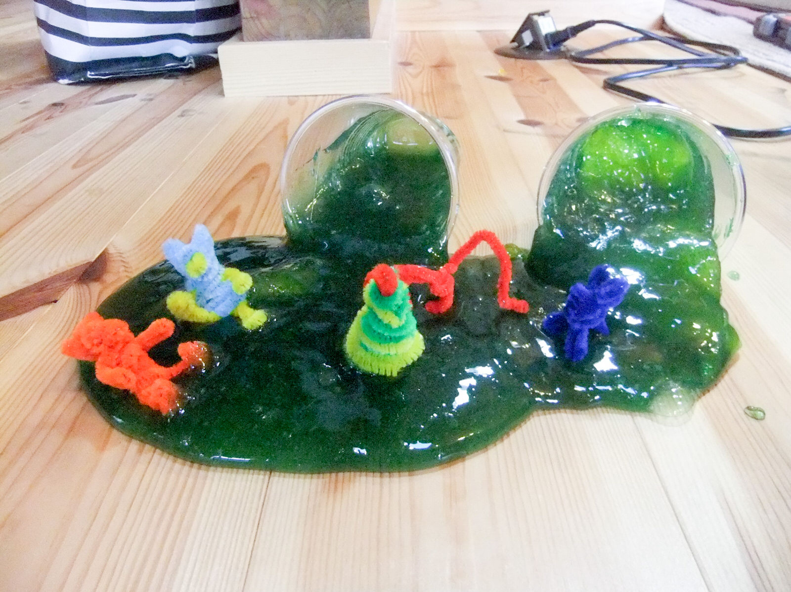 木の床の上に緑色のスライムが入った透明のカップが2つ並んで横倒しに置かれ、そこから床に広がったスライムの中にカラフルなモールで作られた小さな人形などが置かれている写真
