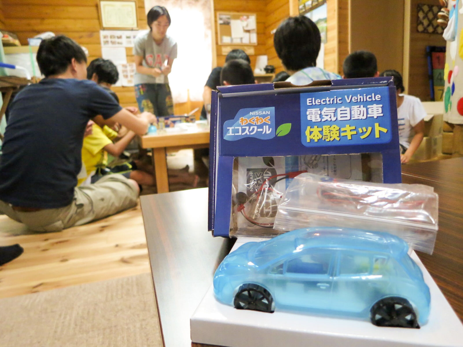 木造の部屋の中で子どもたちと大人が座卓を囲んで座り工作に取り組んでおり、手前には「電気自動車体験キット」と書かれた箱と半透明の水色のおもちゃの車が置かれている写真